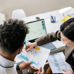 Two colleagues in a meeting room discussing financial charts and graphs on a laptop and paper.